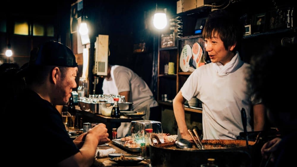 a japanese man in a restaurant is smiling and chatting with a customer at the bar