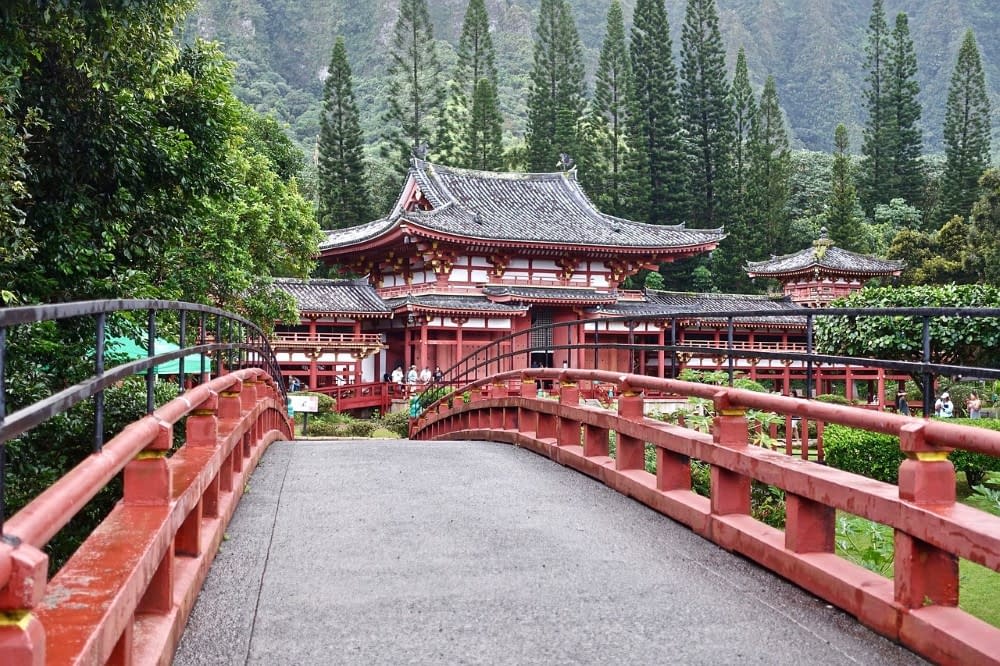 A traditional Japanese bridge, with some traditional Japanese buildings in the background, the bridge is painted red to match the buildings and there are hills and trees behind them