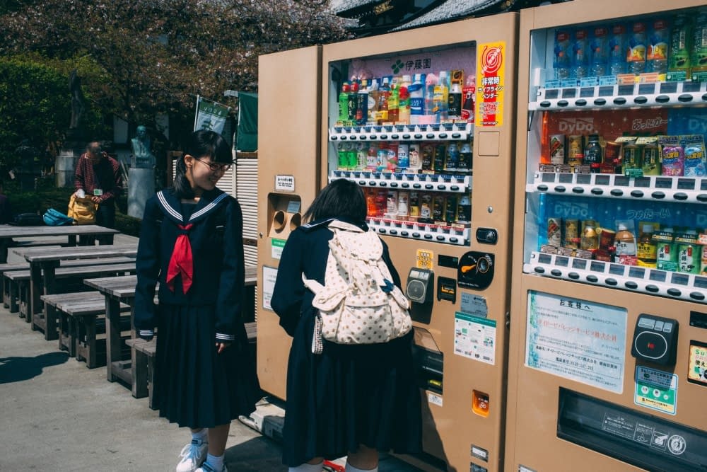 Two Japanese students standing by a vending machine, choosing their favorite snacks.
