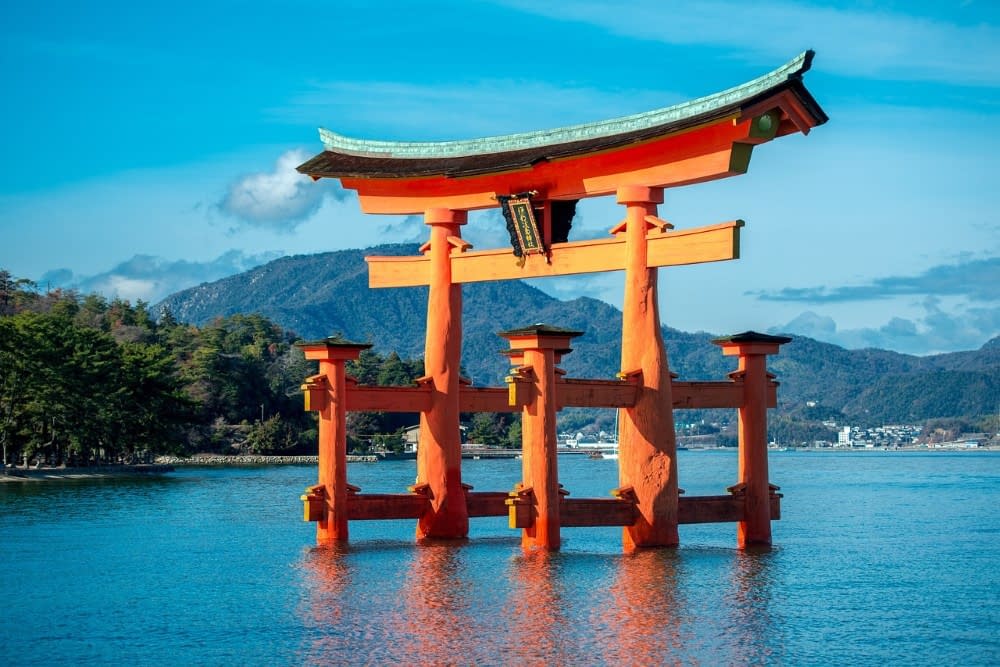 A Japanese Torii Gate in a body of water, with blue sky and mountains in the background