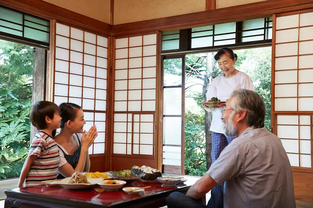 A Japanese family are seated on the floor around a table, as an older woman serves traditional food