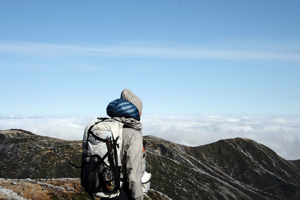 A hiker looks looks out across rolling clouds and the expanse of the horizon at the summit
