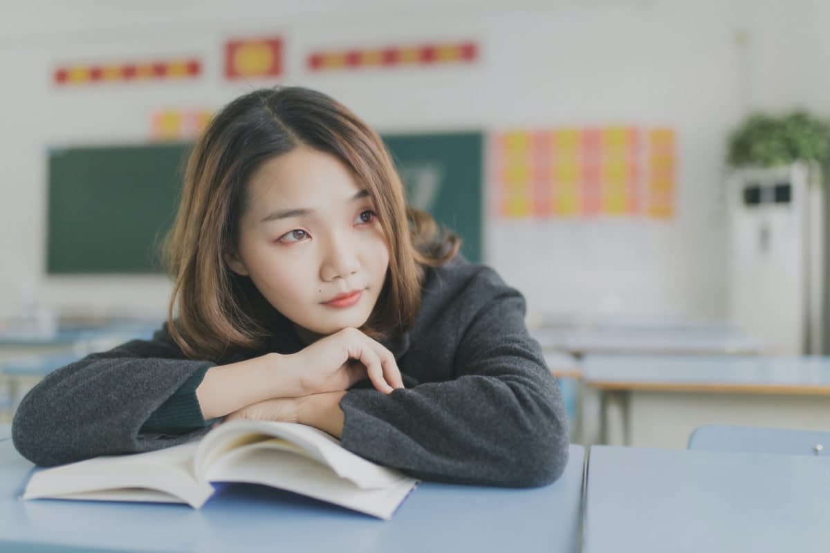 A Japanese woman in a classroom resting on a book and staring off into the distance