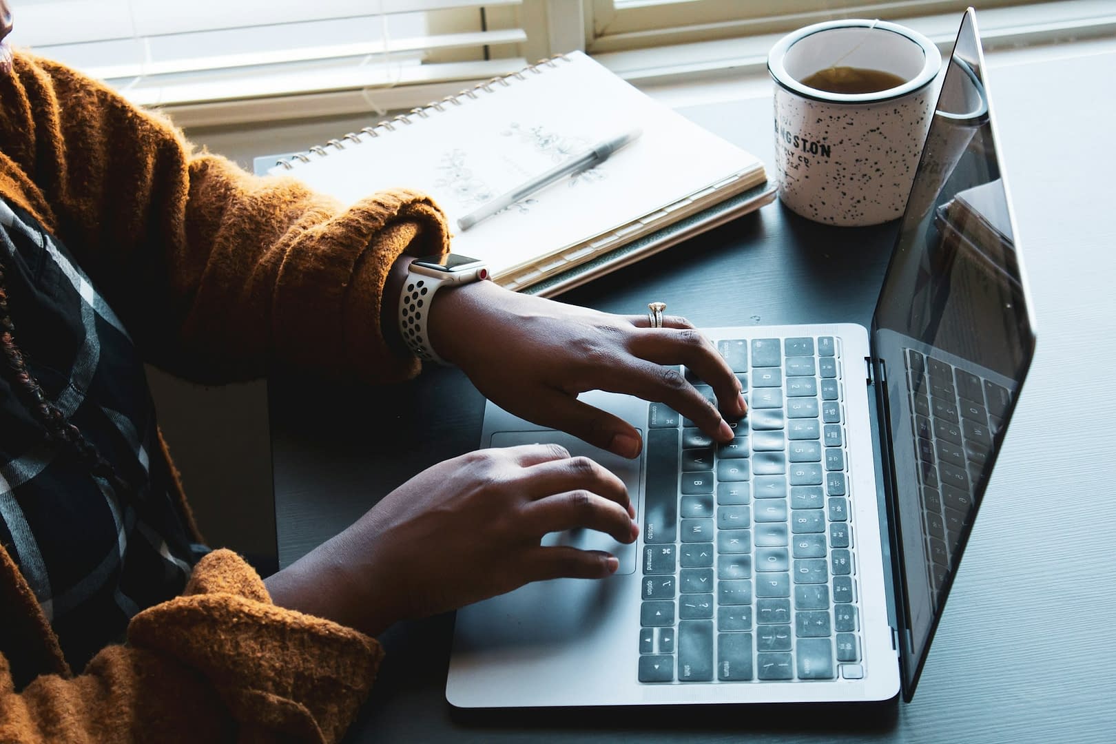 A woman sat at a table, typing on a laptop, with a notebook and cup of coffee beside her.
