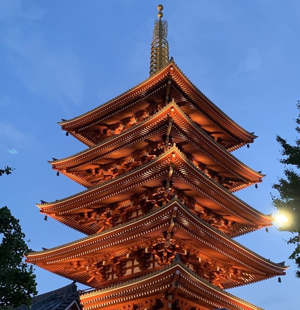 A tall red traditional Japanese tower illuminated by lights, with a blue sky in the background and some trees in the foreground