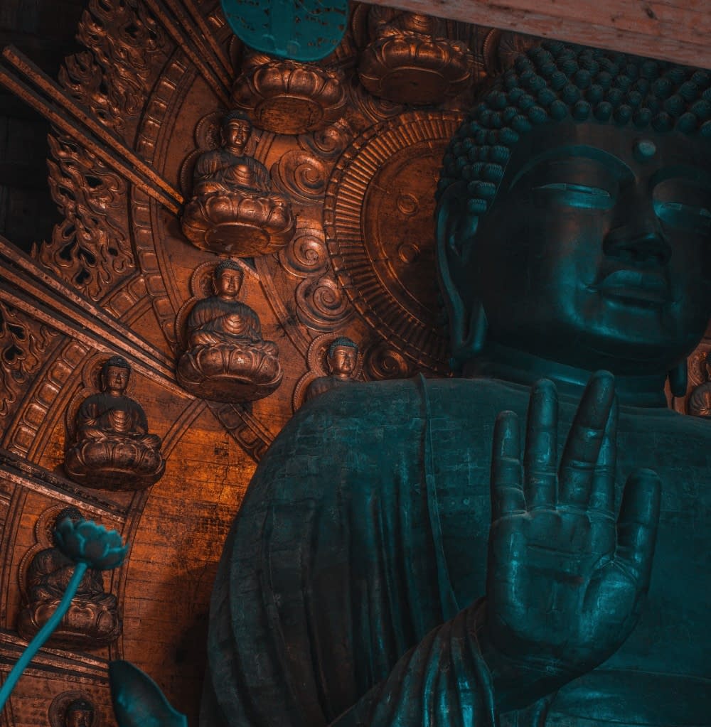 A close up of a Japanese Buddha statue with its hand out, against a metal backdrop with various smaller Buddha statues