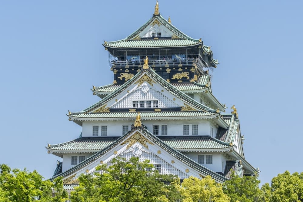 A white and gold traditional Japanese building with a blue sky in the background and green trees in the foreground