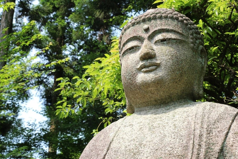 A close-up image of a stone Japanese Buddha statue with trees in the background