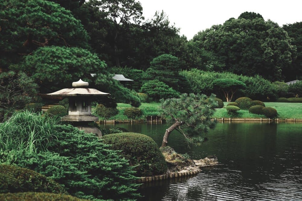 A small shrine sitting by the lake of a quiet, vividly green Japanese garden.