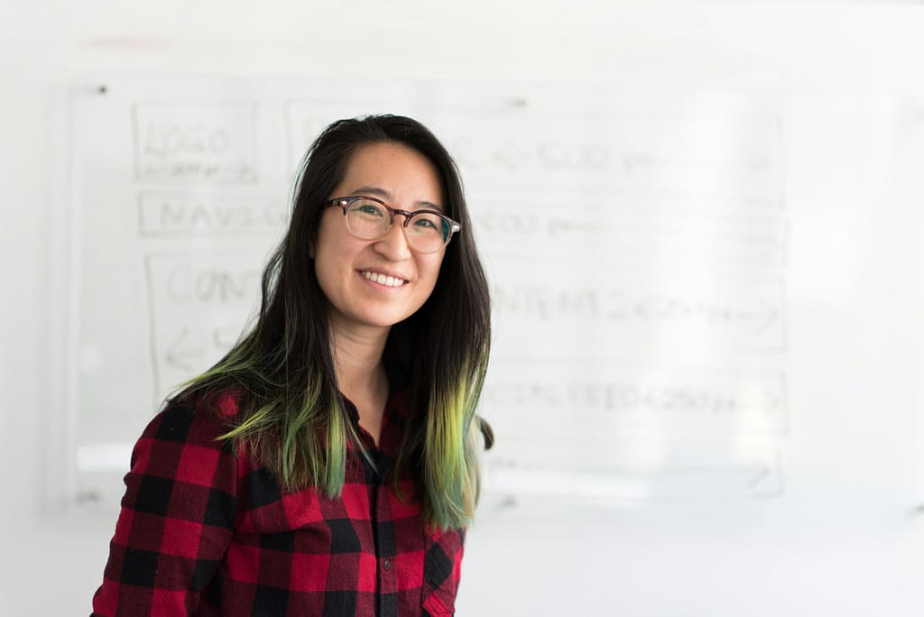 A smilng East Asian woman with black hair and green highlights is smiling in front of a whiteboard with writing on.